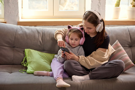 Mother and 5 years old daughter sitting on couch in living room, wearing headphones and looking at smartphone, playing, watching videos.の写真素材