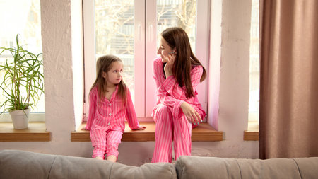 Mother and daughter spending time together at home, sitting on windowsill in matching pink pajamas...