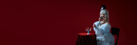 Dramatic scene. Woman in historical image raising opera glasses above red table and expecting pomegranates against deep red backgroundの写真素材