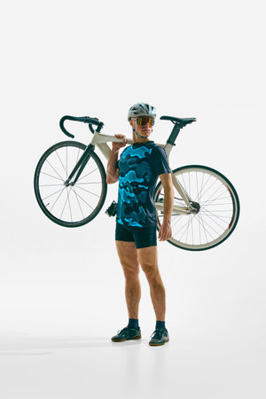 Full-length image of serious young man, cyclist in uniform, helmet and goggle holding bicycle on shoulder isolated on white background.の写真素材