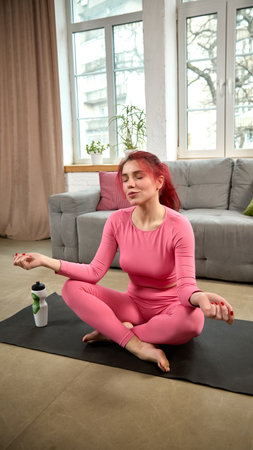 Young woman in pink sportswear training at home in living room, sitting in lotus pose, meditating on yoga mat with peaceful expression.の写真素材