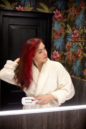 Woman with red hair, in bathrobe brushing hair looking in mirror in bathroom at home. Morning routine, grooming.の写真素材