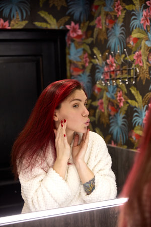 Woman in bathrobe makes kissy face in mirror, touching cheek, doing everyday skincare routine at home in modern bathroom.の写真素材
