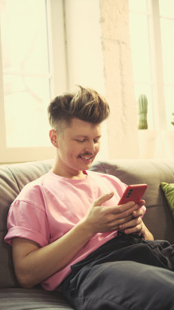 Happy man in pink shirt smiling, using smartphone, sitting on couch in cozy living room at home in daylight.の写真素材
