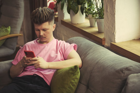 Focused man with smartphone sitting by bright window on comfortable sofa at homeの写真素材