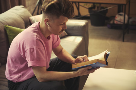 Man with wireless earbuds reading book on modern sofa in sunlight at home in cozy living room.の写真素材