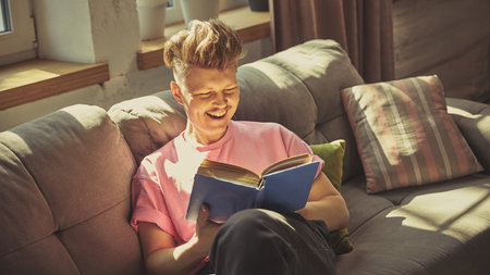 Man in pink shirt smiles joyfully while reading blue book on cozy sofa under daylight in living roomの写真素材