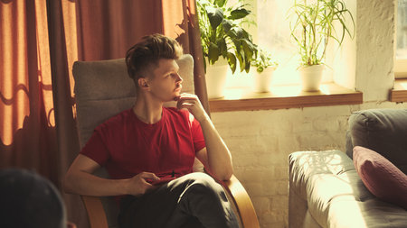 Man in red shirt sits in armchair looking thoughtful with hand on chin. Resting at home in living room.の写真素材