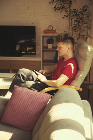 Man in red shirt with smartphone sits in stylish armchair, looking at TV, modern interior of living room.の写真素材