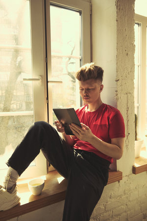 Man in red shirt sitting on windowsill with cup of tea, reading on digital tablet in sunlight at home.の写真素材