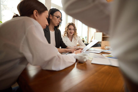 Three women, colleagues look at documents, discuss work at wooden table in modern office, pointing at papers.の写真素材
