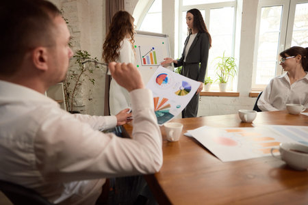 Colleagues discuss charts and data during presentation near whiteboard in bright office, gesturing toward visuals.の写真素材