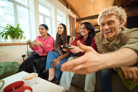Cheerful friends playing video games on couch and enjoying tea together in casual loft interior.の写真素材