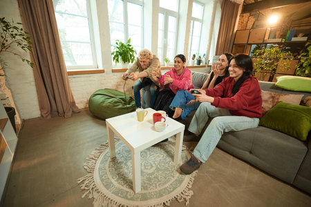 Joyful friends playing video games on couch and enjoying tea together in modern living room interiorの写真素材