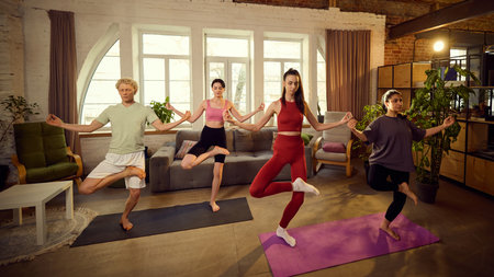 Group of friends practicing Tree Pose in yoga, balancing on one leg with hands in prayer position, standing on yoga mats in spacious room.の写真素材
