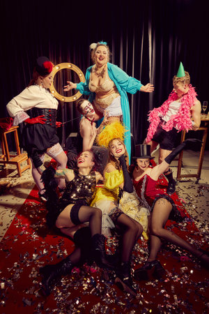 Men and women in eccentric, vintage-style outfits pose dramatically on confetti-strewn red carpet,...