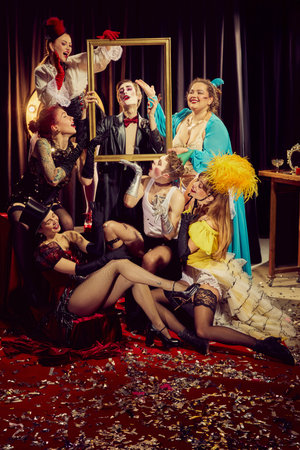 Group of expressive cabaret artists, adorned in vintage clothing, playfully interact with gold frame, posing on confetti-covered red carpet.の写真素材