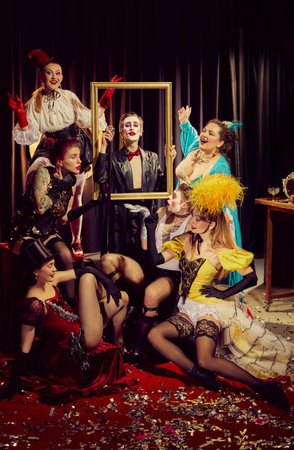 Cabaret performers in mix of dramatic and whimsical vintage costumes gather around gold frame on red carpet covered in confetti.の写真素材