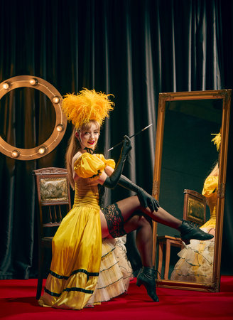 Female elegant performer, dressed in yellow vintage style outfit, seated elegantly on chair on red carpet, posing with cigarette holder.の写真素材