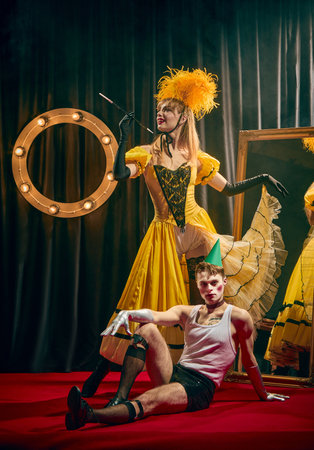 Two cabaret artists, in dramatic yellow vintage dress and in expressive clown-like makeup, pose together on red carpet with confettiの写真素材