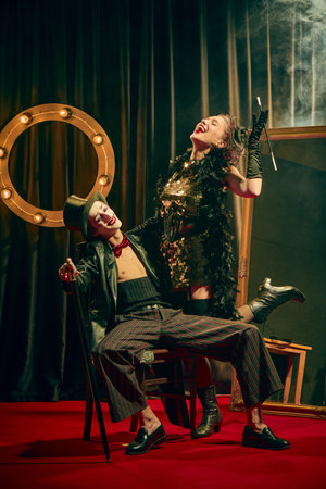 Expressive laughter and dramatic poses of two cabaret performers in vintage costumes on confetti-covered red carpet with smoke, lively moment.の写真素材