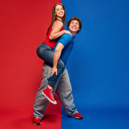 Full-length of young man giving young woman a piggyback ride, both looking up and smiling, against vivid red and blue background.の写真素材