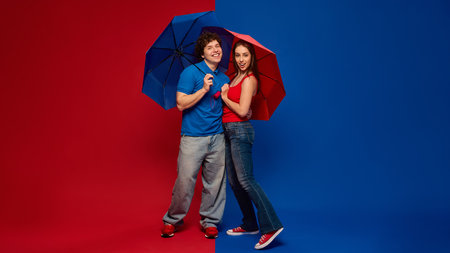 Cheerful young man and woman, couple standing under red and blue umbrellas and smiling at camera, against vivid red and blue background.の写真素材