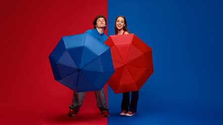 Playful young man and woman, couple posing with large red and blue umbrellas and smiling against vivid red and blue background.の写真素材