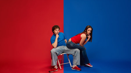 Thoughtful young man and woman, couple sitting together on stool with pensive expressions, against vivid red and blue background.の写真素材