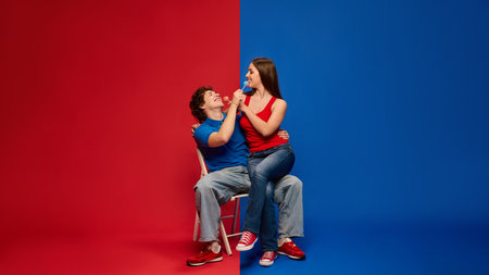 Cheerful young man and woman, couple seated together on stool holding lollipops and smiling at each other, against vivid red and blue background.の写真素材