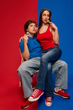 Thoughtful young man and woman, couple seated together on stool with lollipops and looking at cameraの写真素材