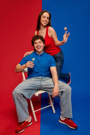 Cheerful young man and woman, couple seated on stool with woman standing behind, holding lollipops and smiling at cameraの写真素材