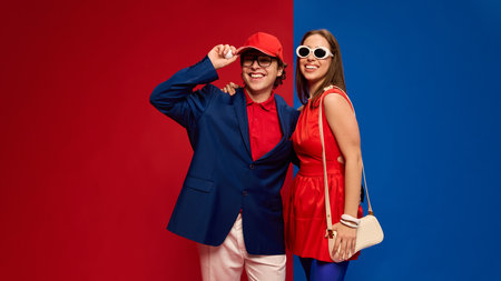 Couple smiling with bright outfits posing together confidently, man tipping cap, holding ball on red blue background.の写真素材