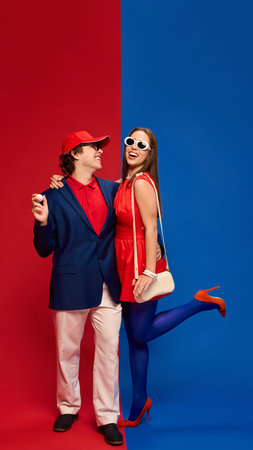 Cheerful young man and woman, couple in golf-inspired outfits, smiling and embracing against vivid red and blue background.の写真素材