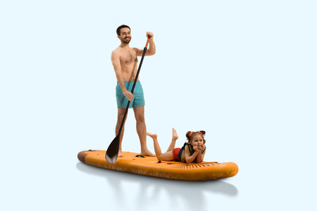 Father smiling and paddling on sup board with his daughter child lying down relaxed in life jacket against neutral studio background.の写真素材
