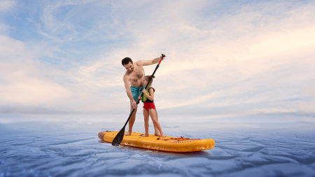 Smiling and caring father paddles on paddleboard, teaching his little daughter to paddle board against blue sky in calm water. Composite image.の写真素材