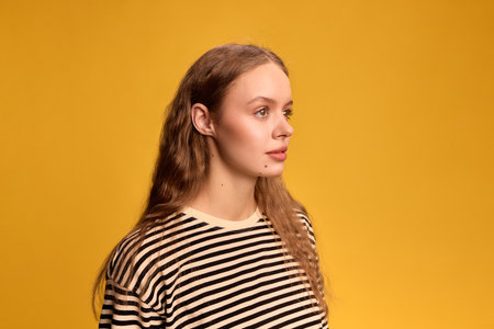 Portrait of young woman with long hair, in striped shirt looking sideways with calm expression against yellow background.の写真素材