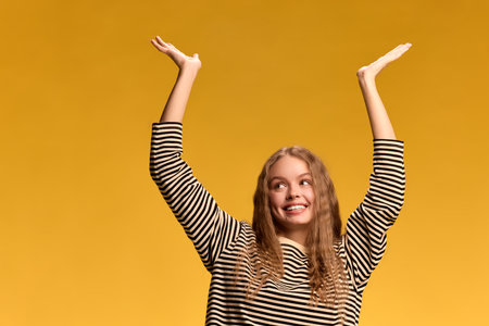 Young woman smiling happily with both arms raised above her head, looking upwards with bright smile against yellow studio background.の写真素材