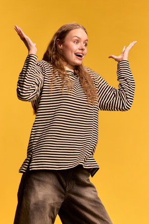 Surprised young woman raising hands expressively with wide eyes and open mouth against yellow background.の写真素材