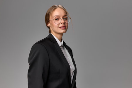 Portrait of smiling businesswoman in black suit and glasses looking aside against grey studio background.の写真素材