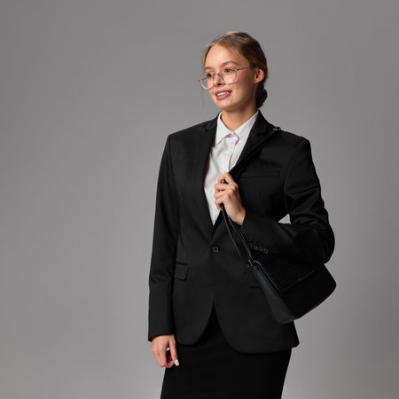 Businesswoman in black suit and glasses smiling gently, holding handbag on shoulder against grey background.の写真素材