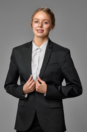 Young beautiful businesswoman in blazer and glasses, posing confidently against grey studio background.の写真素材