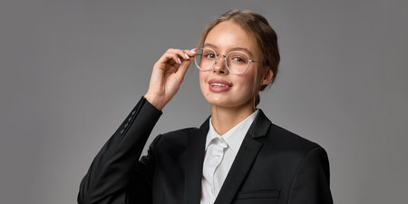 Close-up portrait of young beautiful woman in formal wear and stylish glasses, posing confidently with smile against grey studio background.の写真素材