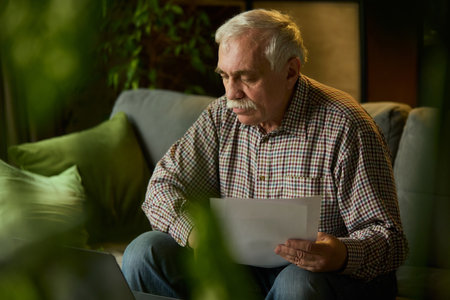 Focused elderly man with mustache sitting on sofa, looking down at papers, studying. Cozy living room with plants.の写真素材