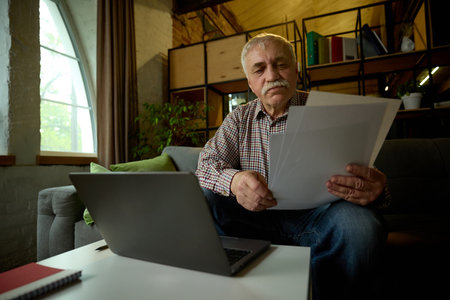 Focused elderly man with mustache sitting on sofa in modern living room, looking at papers, with laptop on tableの写真素材