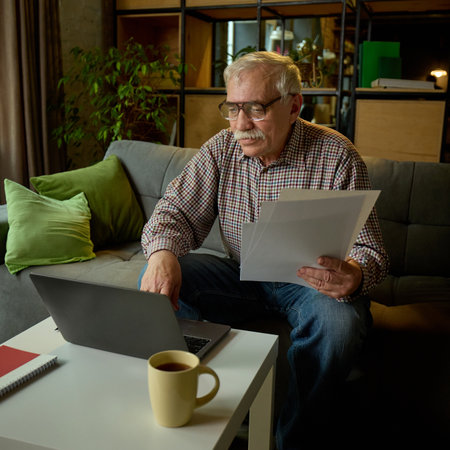 Concentrated elderly man in glasses sitting on sofa in modern living room, looking at laptop and holding papers, studying online with tutor.の写真素材