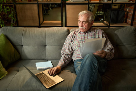 Senior man sitting on grey sofa, using laptop and holding documents in relaxed home environment. Self-development, active aging.の写真素材