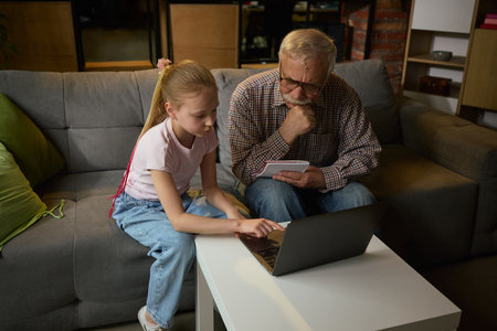 Young girl helping elderly man with laptop while studying together on sofa in cozy home setting. Two generations, support and development.の写真素材