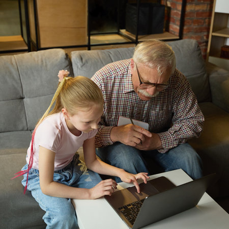 Child girl using laptop while elderly man observing with attention and taking notes during home learning sessionの写真素材