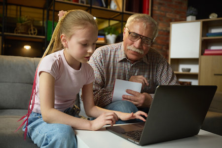 Child girl focusing on laptop while elderly man observing with attention and taking notes during home learning session.の写真素材